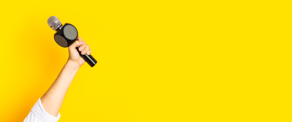 Man's hand holds a microphone in a hole in a torn bright yellow cardboard background. Banner