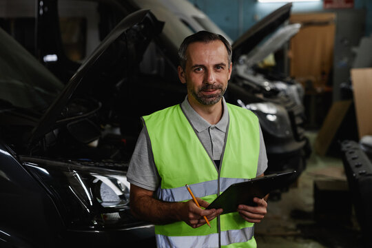 Waist up portrait of mature male worker looking at camera while inspecting vehicles at car factory quality control