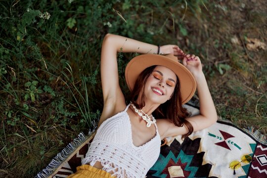 A woman in eco-friendly clothing in a hippie look is lying on a colored plaid smiling and looking at an autumn sunset in nature in the park. Lifestyle on a journey of harmony with the world