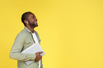 Laughing adult african american guy in wireless headphones with laptop looking up at empty space