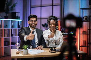 Diverse colleagues, smiling successful african american businesswoman and indian it specialist in formal wear recording new video for their internet business course showing thumb up.
