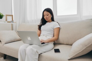 Asian woman sitting with laptop and phone in freelancer's home office looking at laptop screen with headphones working, lifestyle work and home