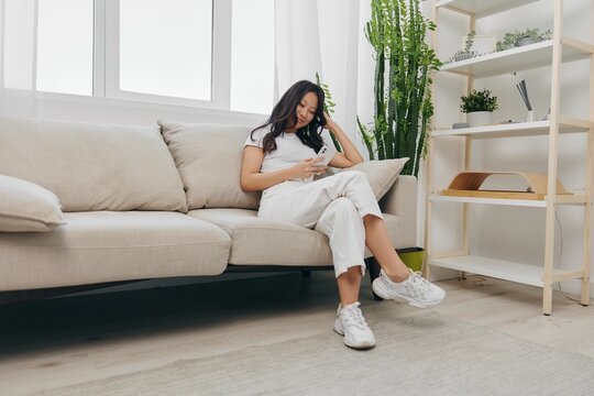 A young Asian woman sits at home on the couch relaxing and flipping through her social media feed and reading messages on her phone blogger home clothes. Lifestyle without work in a comfortable home