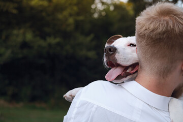 Dog owner hugs his young dog outdoors. Man interacting with a white staffordshire terrier puppy, happiness, joy and positive emotions with pets