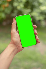 La mano del hombre joven y delgado  sosteniendo un smartphone móvil con pantalla verde en posición vertical en fondo de un jardin. Maquetear móvil
