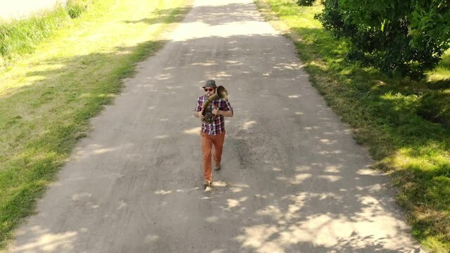 A jolly trumpeter with glasses and a hat walks and plays the trumpet on the road in the park. Drone view from above