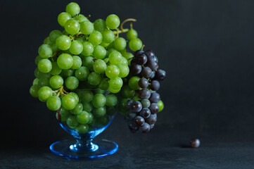 Clusters of black and green grapes in a glass bowl on a black shabby background. Selective focus with copy space. Daylight