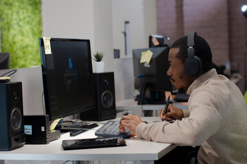 African American male artist designer in headphones draws on tablet 3D models for games. Against background, multinational team of developers are discussing concept of presentation of project.