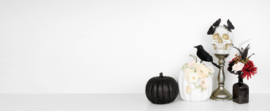 Elegant Halloween shelf decor with copy space against a white wall banner background. White shelf with red and black flowers, skull with butterflies, pumpkins and crow.