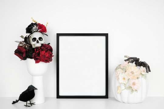 Halloween elegant gothic mock up. Black frame on a white shelf with red and black flowers, skull and floral pumpkin. Portrait frame against a white wall. Copy space.