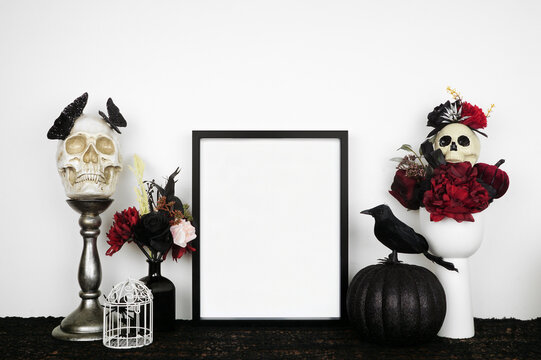 Halloween gothic romance mock up. Black frame on a black shelf with red and black flowers, skulls and pumpkin. Portrait frame against a white wall. Copy space.