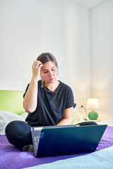 Vertical shot of Caucasian pensive woman looking at computer from bedroom