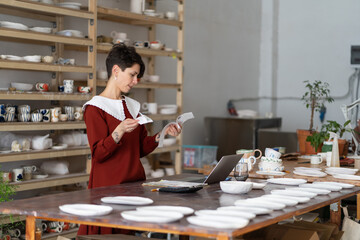Focused female ceramist looking at receipts, calculating material costs and rent expenses while standing in her pottery studio, craftswoman small business owner thinking how to fix up financial issues