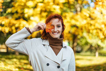 Portrait of glad caucasian millennial red haired female in raincoat puts yellow leaf to her eye