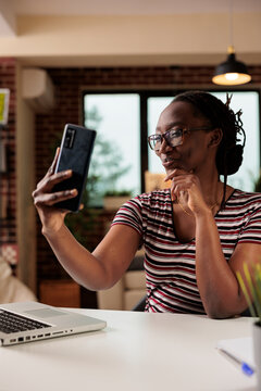 Young woman taking selfie on smartphone, posing, smiling at front camera. Attractive freelancer making portrait photo on mobile phone at home office workplace, remote worker having video chat