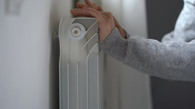 Woman touches room central heating radiator feeling cold in apartment. Lady tries to warm up putting hands in sweatshirt closer to battery and wants to keep warmth in room. Utility services problems