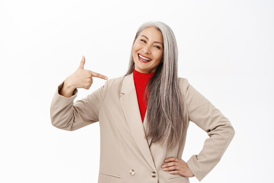 Happy Smiling Stylish Old Woman In Suit, Pointing Finger At Herself And Looking Pleased, Self-satisfied, Standing Over White Background