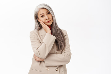 Portrait of stylish beautiful asian senior woman, lean head on hand, looking with bothered and tired face expression, standing over white background