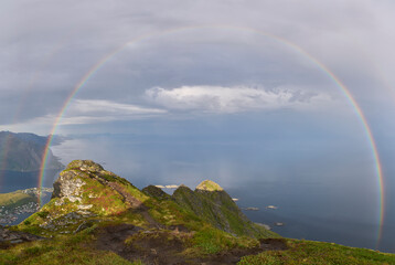Panoramic view on colorful rainbow above sea from Reinebringen ridge, Lofoten, on summer day. Inclement weather, storm clouds. Scenic vista with a rainbow and rugged peaks. Vacation in Lofoten.