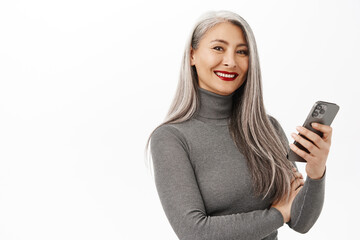 Stylish asian senior woman, beautiful lady holding mobile phone, posing against white background with happy smiling face expression