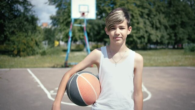 Portrait of a boy with a ball on a basketball field looking at the camera and smiling, moving camera.