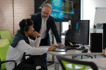 Caucasian CEO of the company studio developers of games and applications discusses with Asian artist a 3D model on a computer against the background of other employees of the company.
