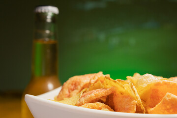 A white plate with chips next to a bottle of beer out of focus with the projection on a wall of a soccer game out of focus in the background