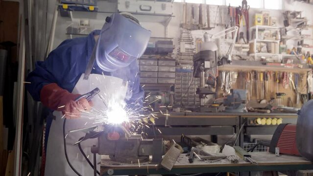 Craftsman working with arc welding machine in workshop. Piece of iron with sparks fly in different directions. Male worker hand welding steel rack.