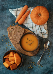 Pumpkin cream soup in a bowl with a spoon on wooden board on the table next to pumpkin whole and cut into pieces.  Healthy, vegetarian food. Diet. Home cooking. Thanksgiving day. Halloween. Top view
