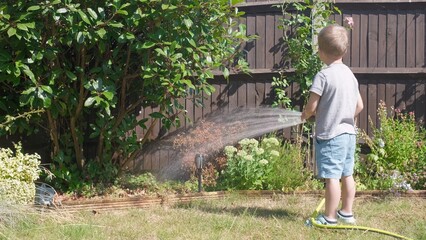 Funny little boy watering lawn plants in garden housing backyard. Adorable child playing with irrigation hose at hot sunny summer outdoors. Children help with housework. activity for kids. Childhood