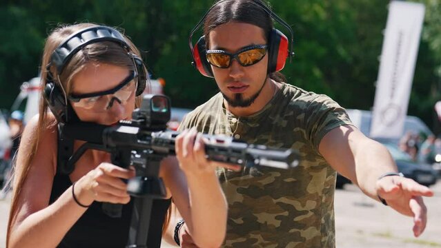 Black rifle with collimator held by a caucasian long-haired girl in a black tank top wearing protective gear during a shooting lesson with her male instructor who is standing at the background