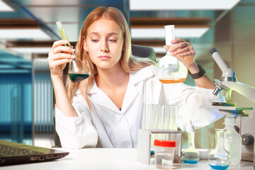 Chemical laboratory assistant. Woman is sits at table with laboratory equipment. Chemist in modern laboratory. Microscope and flasks in front of chemist. Blond woman works as chemist.