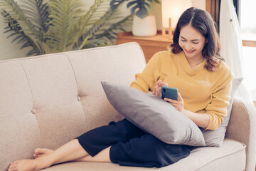 Bright and cute Asian young woman wearing headphones sit on the sofa and using smartphone to listen to music happy and relax listening to music at home.