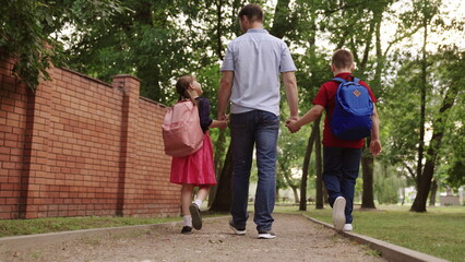 happy family. father holding hands children school. little kids school bags go school holding dad hand. concept happy family outdoors. schoolchildren with parent view from back. children education.