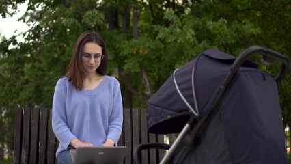A young mother works on a laptop and shakes her baby in a stroller. A woman with glasses works remotely. Woman on a bench in the park.