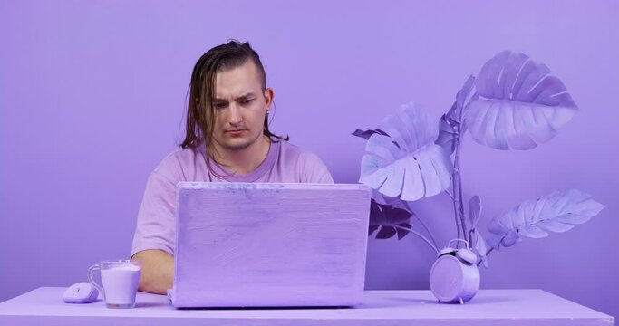 Long-haired young man is sitting at laptop in lilac-colored room and attentively looks at screen. Unusual creative cafes and coworking offices for freelancers. Interior of room in very peri color.