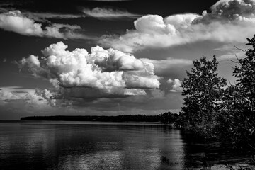 Dramatic black and white image of large white clouds over a calm lake on a bright summer day. 