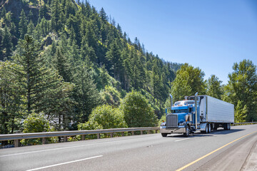 Blue American idol classic bonnet big rig semi truck tractor transporting frozen food in reefer semi trailer driving on the highway road with forest on the side