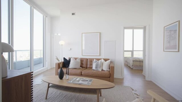 Wide angle orbit shot of the living area of a high ceiling penthouse condominium unit with a floor to ceiling window wall at the side and balcony outside during a sunny day