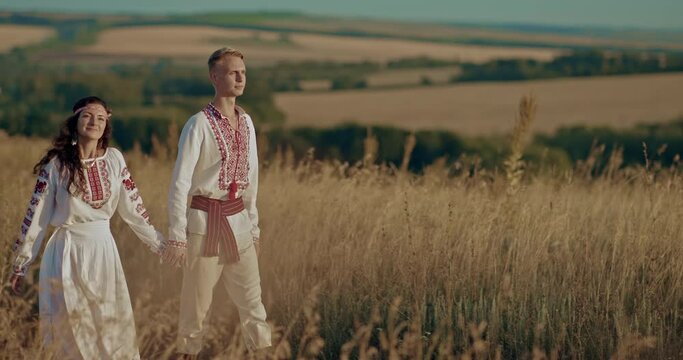 Happy man and woman walk through the village in Slavic costumes and enjoy life. Beautiful couple of men and women in embroidered shirts walking and holding hands, friendship of peoples. 4k, ProRes