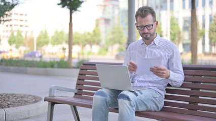 Young Adult Man Reading Documents and Working on Laptop while Sitting on Bench Outdoor