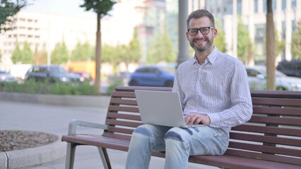 Young Adult Man with Laptop Smiling at Camera while Sitting Outdoor on Bench