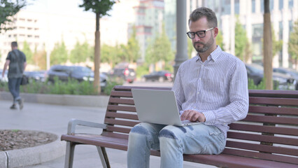 Busy Young Adult Man Using Laptop Sitting Outdoor on Bench