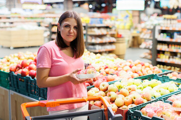 Young brunette woman picks chooses fruits on the counter in supermarket. Female housewife shopping in market standing near vegetable department store with a grocery cart.