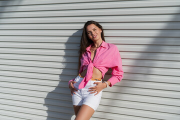 Young beautiful brunette woman in a pink shirt and white denim shorts against the background of a...