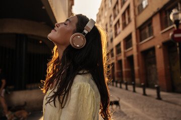 Nice young caucasian girl eyes closed listens to music using headphones standing on street. Brunette is having relaxed time, wearing dress. Audio technology concept