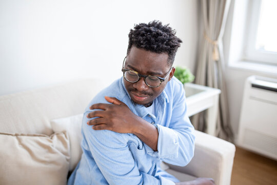 Pain in his shoulder. Upper arm pain, People with body-muscles problem, Healthcare And Medicine concept. Attractive young man sitting on the sofa and holding painful shoulder with another hand.