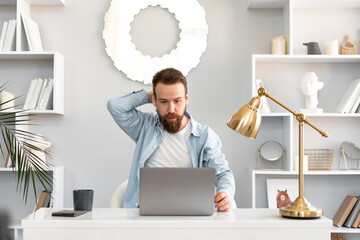 Thoughtful tired young bearded man sitting at the table working on laptop