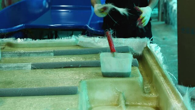 A worker is tearing off a piece of fiberglass to finish the hull of the boat. On the front of the film is a container with glue and a roller. Fiberglass boat construction in a private shop