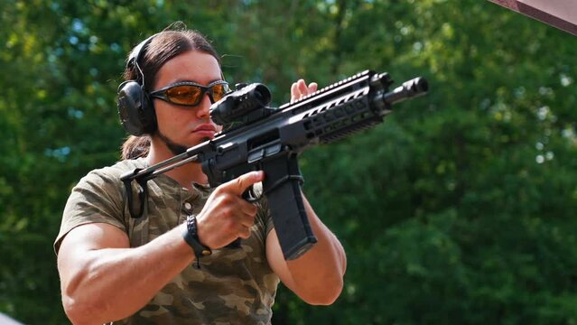 Caucasian bearded man in safety goggles and headphones wearing camo t-shirt reloading submachine gun. Outdoor horizontal shot. High quality 4k footage